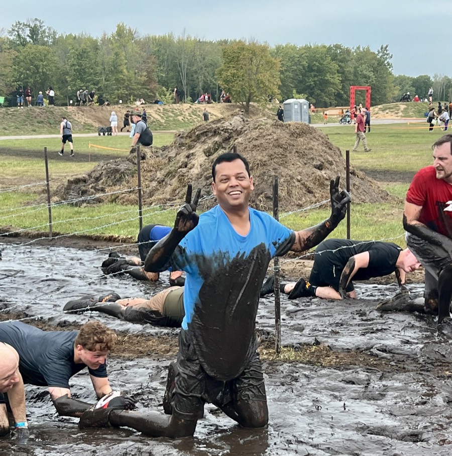 Arvind Choudhary kneeling in deep mud under barbed wire at Tough Mudder, covered in mud and flashing peace signs with both hands.